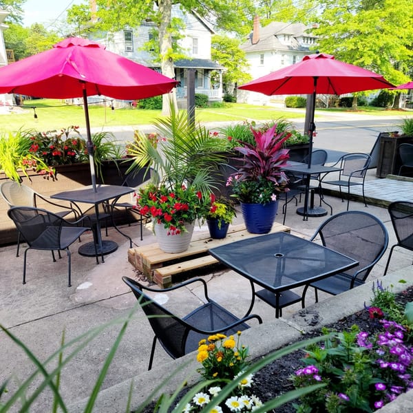 Outdoor patio with black metal tables and chairs, red umbrellas, and vibrant potted plants.