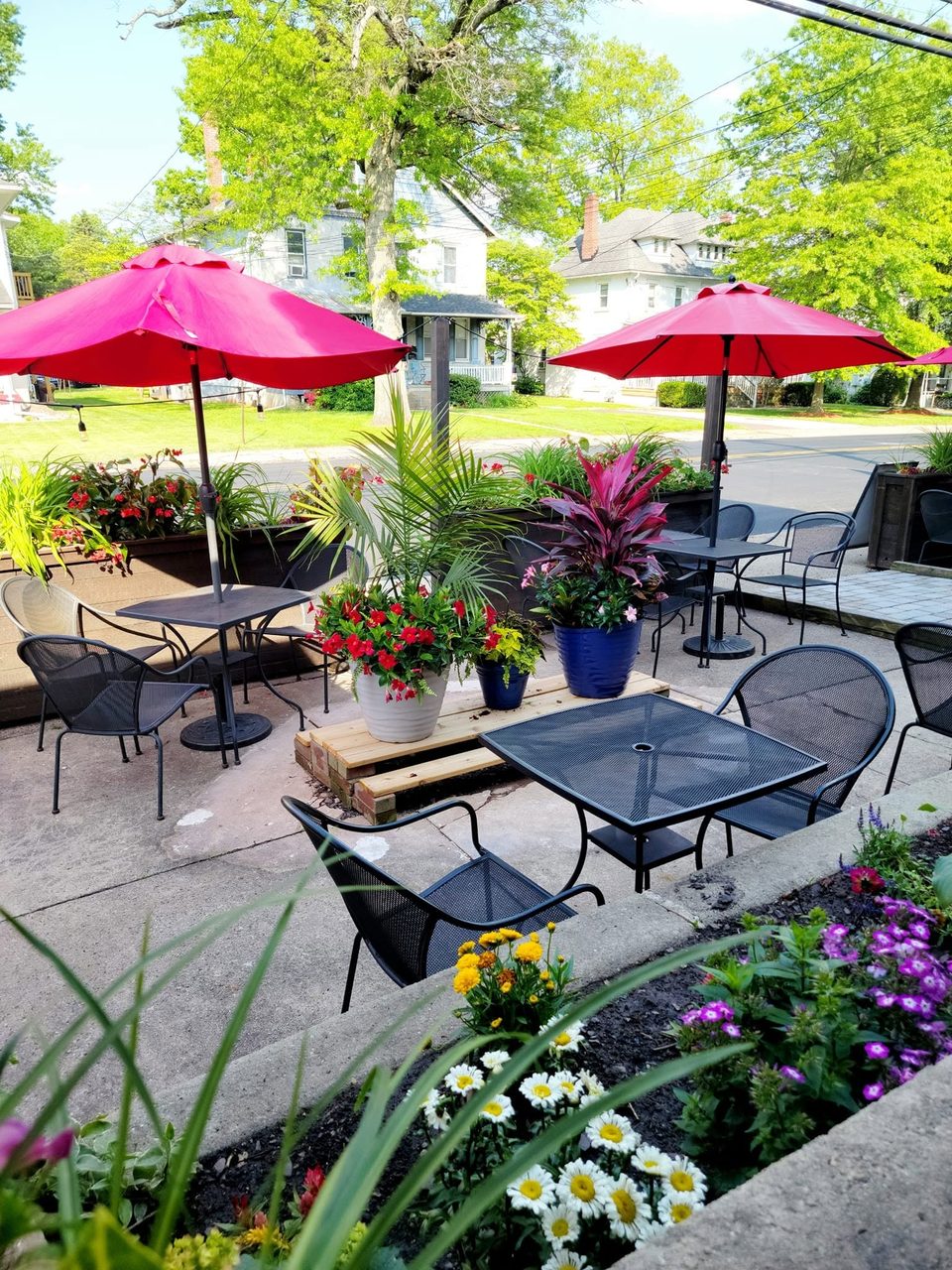 Outdoor patio with black metal tables and chairs, red umbrellas, and vibrant potted plants.