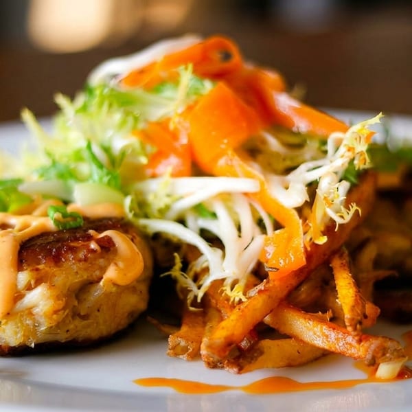 Two crab cakes drizzled with sauce, served with a side of fries and a fresh salad.