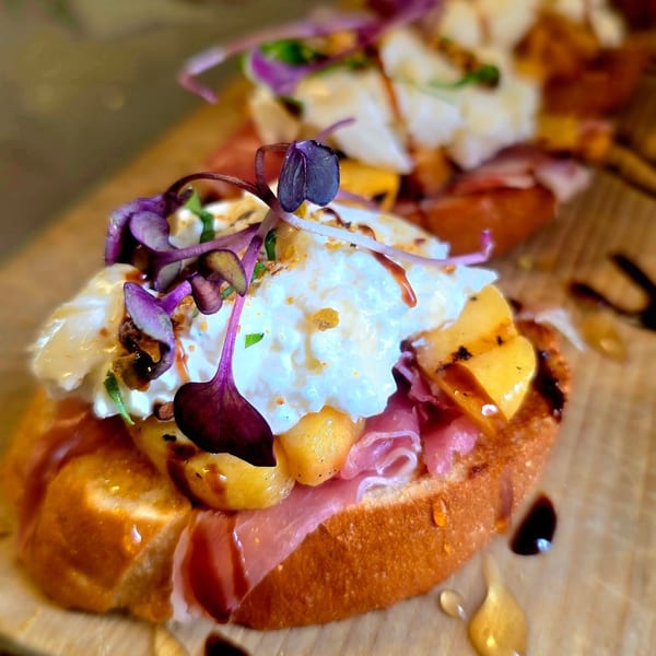 Close-up of bruschetta with prosciutto, peaches, burrata, balsamic glaze, and purple microgreens.
