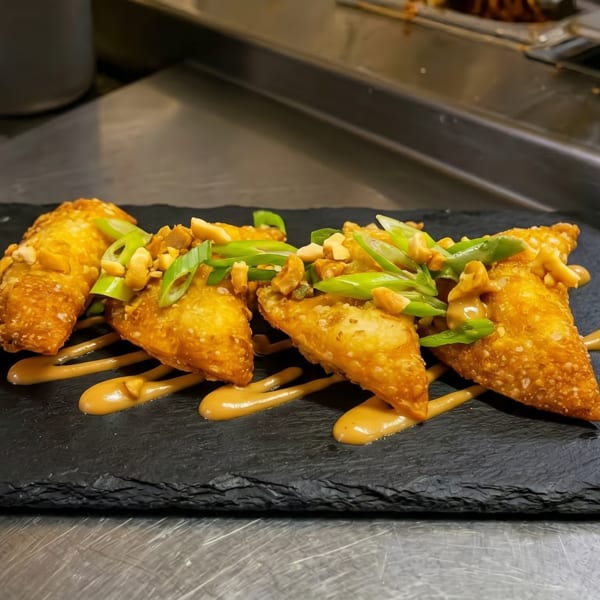 Four crispy fried wontons, drizzled with sauce, topped with peanuts and green onions on a black slate.