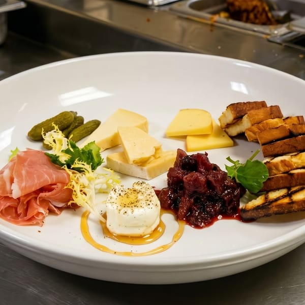 Charcuterie board with prosciutto, cheeses, pickles, jam, goat cheese, and toasted bread on a white plate.