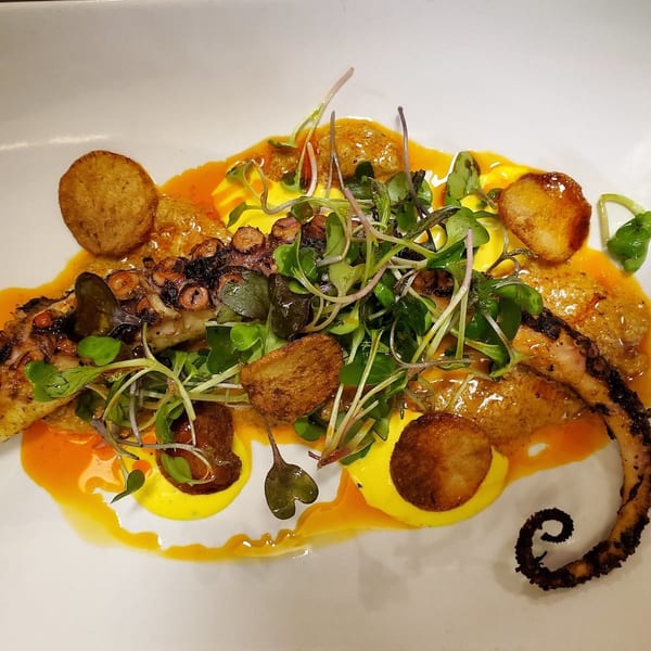 Grilled octopus with microgreens, crispy potatoes, and yellow and orange sauces on a white plate.