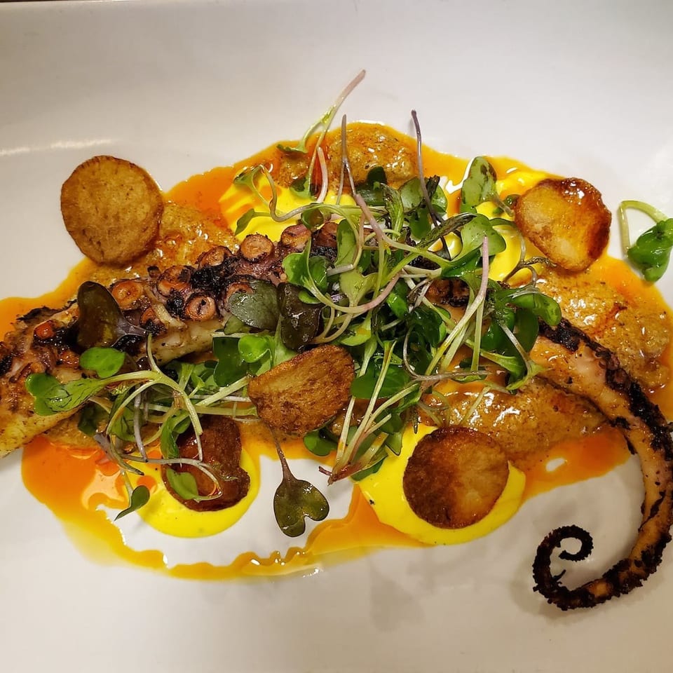 Grilled octopus with microgreens, crispy potatoes, and yellow and orange sauces on a white plate.