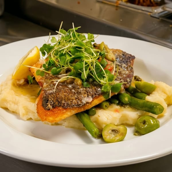 Pan-seared fish with mashed potatoes, green beans, olives, and microgreens on a white plate.