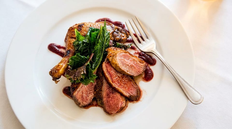 Roasted duck leg and sliced duck breast with greens and berry sauce on a white plate.