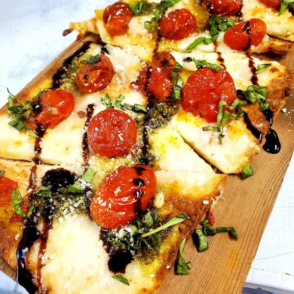 Flatbread pizza with roasted tomatoes, pesto, mozzarella, basil, and balsamic glaze on a wooden board.