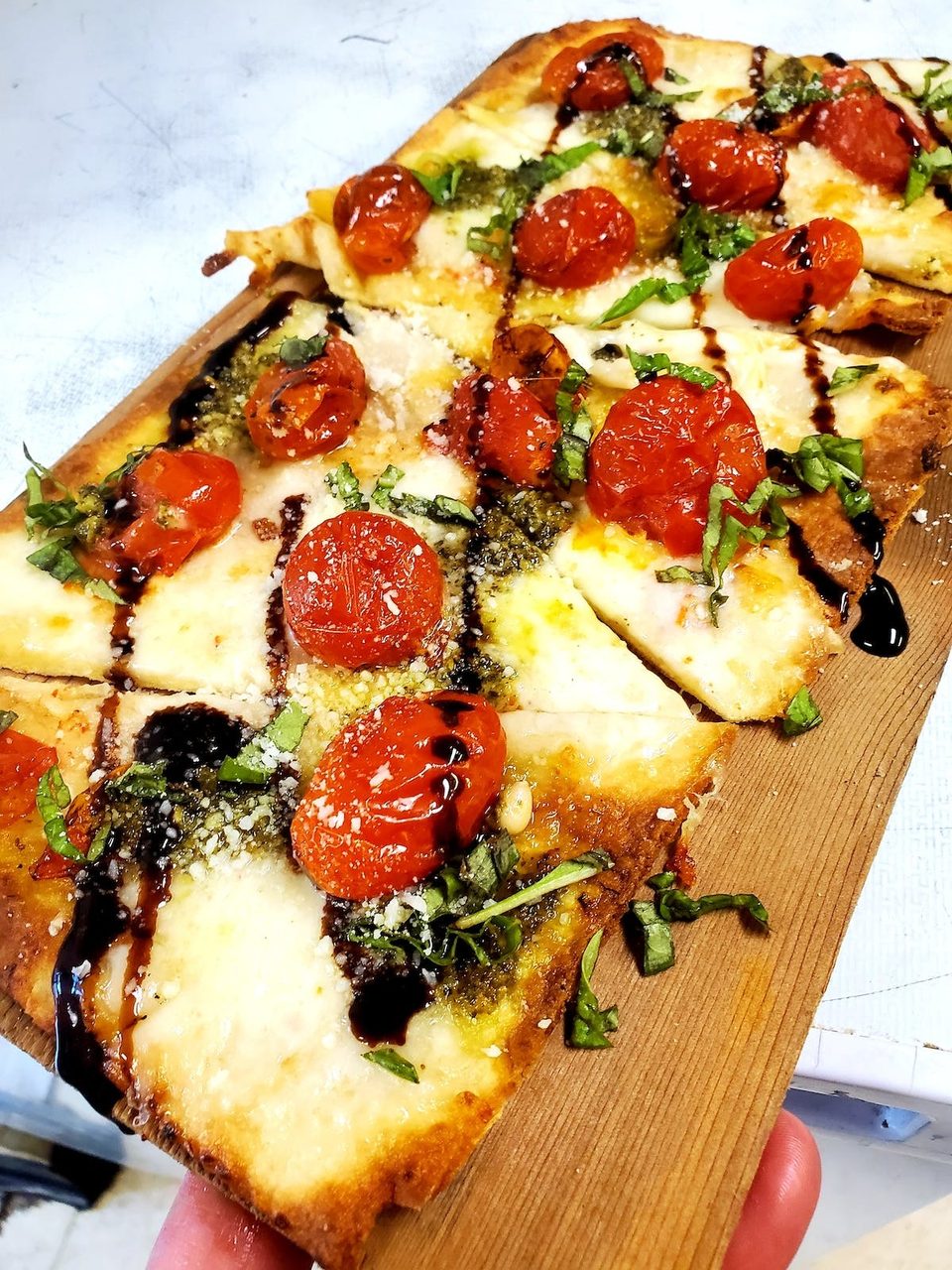 Flatbread pizza with roasted tomatoes, pesto, mozzarella, basil, and balsamic glaze on a wooden board.