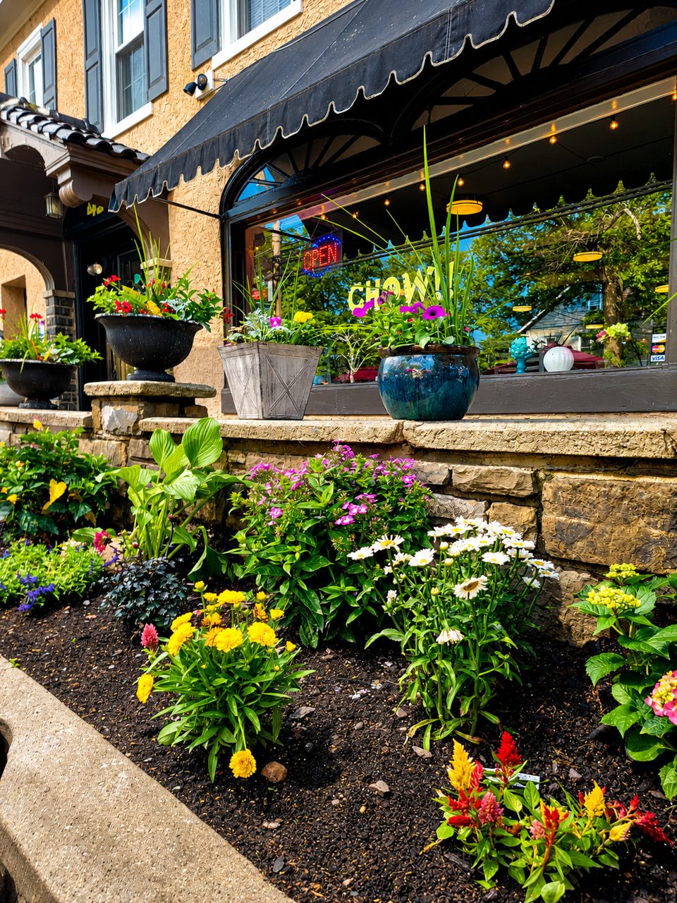 A vibrant flower bed and potted plants adorn the exterior of a restaurant with a black awning.