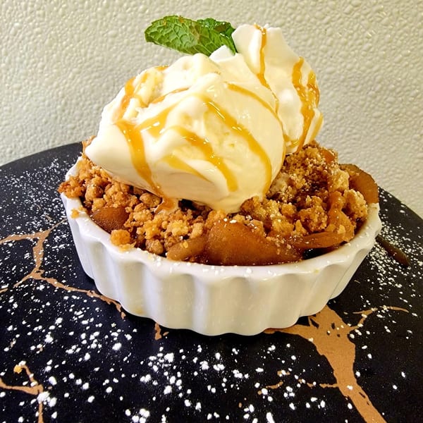 Apple crumble dessert with vanilla ice cream, caramel drizzle, and mint on a decorative plate.