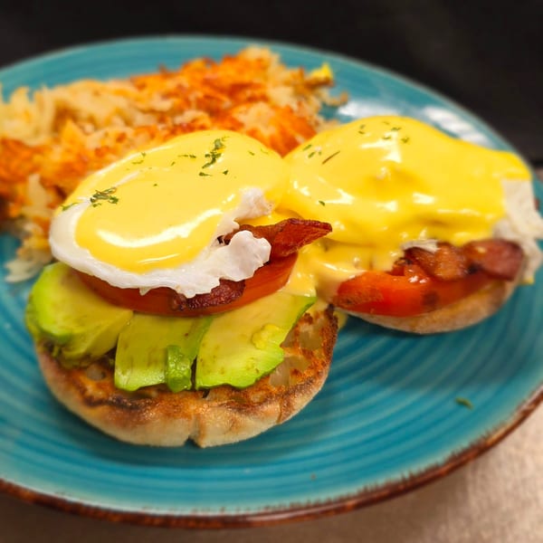 Two eggs benedict with avocado, tomato, and bacon on a blue plate with hash browns.