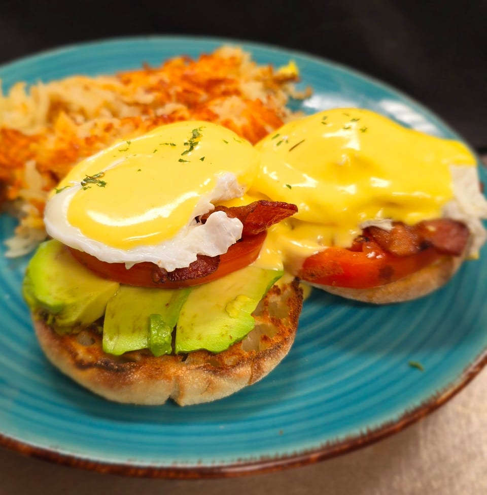 Two eggs benedict with avocado, tomato, and bacon on a blue plate with hash browns.