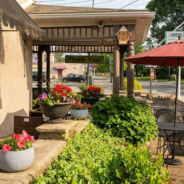 Outdoor patio seating with black tables and chairs under a red umbrella, surrounded by lush plants and flowers.
