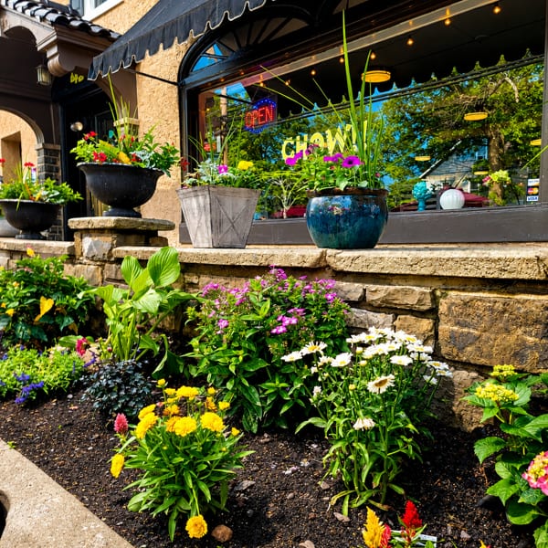 A vibrant flower garden and potted plants adorn the exterior of a restaurant with a black awning.